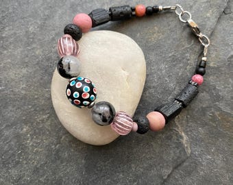 May include: A black and pink beaded bracelet with a silver clasp. The bracelet features black lava rock beads, pink and white wooden beads, and a black bead with white and blue polka dots.