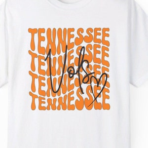 May include: White t-shirt with an orange and black graphic design. The graphic features the word "Tennessee" repeated multiple times in a wavy font, with the word "Vols" written in black script in the center. A small heart is next to the word "Vols".
