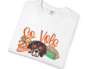 May include: White t-shirt featuring an orange and black graphic. The design includes the words "Go Vols" in orange script, a cartoon dog wearing star-shaped sunglasses, a pom-pom, and a football field.
