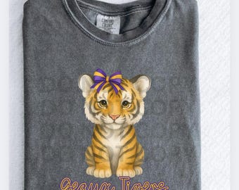 May include: A dark gray Comfort Colors t-shirt featuring a cartoon tiger cub with a purple and gold bow. The tiger has orange and black stripes. The text "Geaux Tigers" is written in a script font below the tiger.