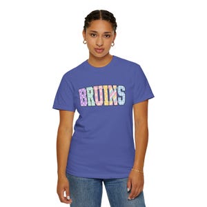 May include: A blue short-sleeved t-shirt featuring the word BRUINS printed across the chest in block letters. Each letter has a unique pastel pattern, including polka dots, floral motifs, and diagonal stripes in shades of pink, yellow, and blue.