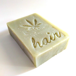 May include: A rectangular, light green bar of soap with the word "hair" carved into the surface. A cannabis leaf design is also carved into the soap. The soap is on a white background.