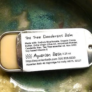 Puede incluir: Un recipiente rectangular de metal plateado que contiene un bálsamo desodorante amarillo claro. La etiqueta dice "Tea Tree Deodorant Balm" y enumera los ingredientes. El producto es de Aquarian Bath y pesa 7 gramos.
