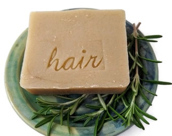 May include: A bar of natural hair soap with the word "hair" embossed on it, resting on a blue and green ceramic dish with sprigs of rosemary.