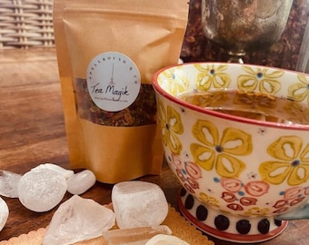 May include: A brown paper bag labeled "Spellbound & Co. Tea Magik" next to a floral teacup filled with tea. Clear crystals and a white candle are arranged on a tan cloth. A silver chalice and a jar of dried herbs are in the background.
