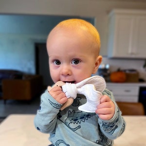 May include: A baby wearing a light blue dinosaur-patterned shirt and blue jeans. The baby is holding a wooden and white fabric teether in their mouth. The teether has a wooden ring and a white, crinkled fabric bow.