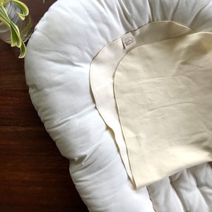 May include: A white, round,  cushioned pillow with a cream-colored,  removable,  fitted sheet. The sheet has a small label that says "Sleepy Jane".
