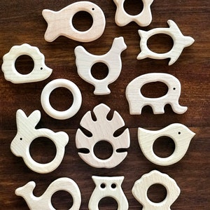 May include: A set of 14 wooden teethers shaped like various animals and objects, including a fish, turtle, chicken, elephant, rabbit, monstera leaf, bird, whale, owl, and a shell. The teethers are all natural wood colour and have a smooth, polished finish.