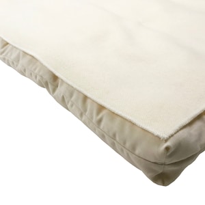 May include: Close-up of a rectangular, off-white cushion with a textured, cream-colored top layer. The edges are rounded, and the fabric appears to be a soft, natural material. The cushion is likely for home decor or comfort.