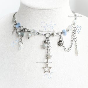 Puede incluir: Un collar plateado con abalorios celestiales. El collar presenta pequeñas estrellas, cuentas y detalles colgantes, incluido un colgante de estrella. Se intercalan cuentas de color azul claro y transparentes. El collar se exhibe sobre una superficie blanca.