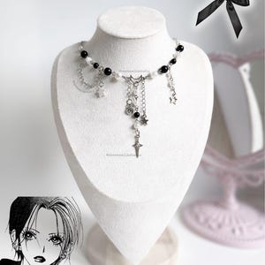 Può includere: Una collana color argento con perline nere e bianche, ciondoli a forma di stella e un ciondolo a mezzaluna è esposta su un manichino bianco. Sono visibili anche un fiocco nero e un'illustrazione in stile manga di una donna.