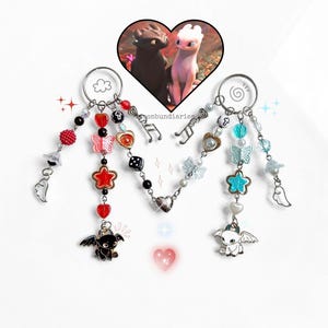 May include: Two keyrings with charms, featuring a heart-shaped image of two dragons. One keyring has red, black, and silver charms, including a dragon, star, and butterfly. The other has blue, white, and silver charms, including a dragon, star, and butterfly.