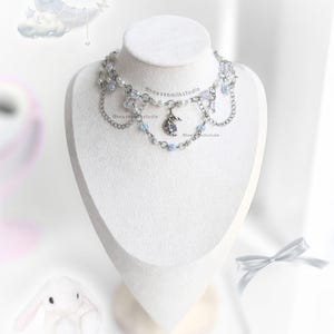 Puede incluir: Un collar plateado con un colgante de media luna, perlas y cuentas azules se exhibe en un maniquí blanco. El collar tiene múltiples capas y detalles de cadena. El texto "heavenmilkstudio" es visible.