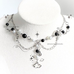 May include: A silver-toned chain necklace featuring black and white beads, small star charms, a heart-shaped charm, and a central cloud pendant. The design includes multiple draped chain layers and the text @heavenmilkstudio printed on the image.