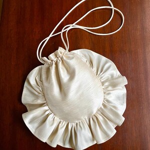 May include: A cream-colored drawstring bag with a ruffled edge. The bag is made of a silky fabric and has a long, thin strap.