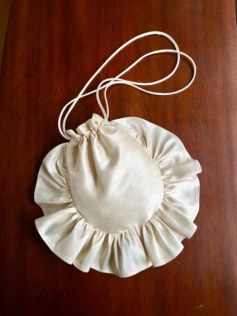 May include: A cream-colored drawstring bag with a ruffled edge. The bag is made of a silky fabric and has a long, thin strap.