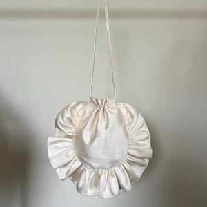 May include: A white, round, ruffled drawstring bag with a long white strap.