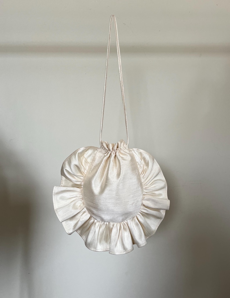 May include: A white, round, ruffled drawstring bag with a long white strap.