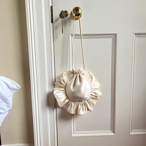 May include: A cream-colored, round, ruffled bag with a drawstring closure and a long, cream-colored strap.