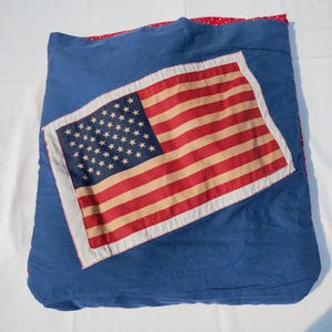 May include: A blue pillow with a large American flag design. The flag features red and tan stripes with a blue square filled with white stars. The pillow has a red polka dot border.