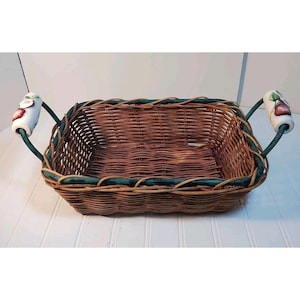 May include: A rectangular wicker basket with a brown woven design. The basket has green trim and white handles with a painted apple design. The handles are attached with green metal. The basket is empty.