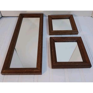 May include: A set of three mirrors with dark brown wooden frames. One mirror is rectangular and elongated, while the other two are square and of different sizes. The frames have a visible wood grain texture and a simple, flat profile design.