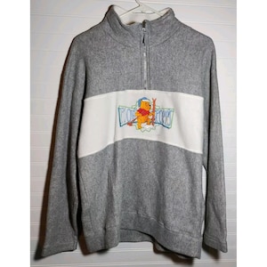 May include: Gray fleece pullover with a white horizontal stripe across the chest. The stripe features an embroidered graphic of Winnie the Pooh on skis with the text "POOH SPORTS". The pullover has a quarter-zip closure and a mock neck.