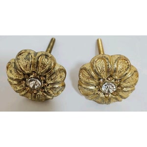 May include: Two antique-style cabinet knobs. Each knob is a golden-colored flower shape with a clear, round crystal in the center. The knobs have a distressed finish and a threaded post for mounting.