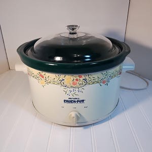 May include: A cream-colored Rival Crock-Pot stoneware slow cooker with a dark green rim and a clear glass lid. The exterior features a decorative fruit and vine border pattern. It has a front-facing control knob with Off, Low, and High settings.