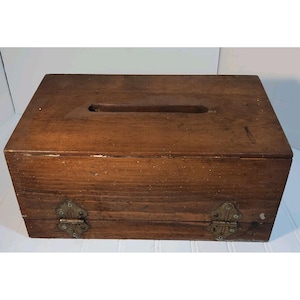 May include: A rectangular wooden box with a dark brown finish and a horizontal slot cut into the top lid. The front panel features two ornate, brass-colored metal hinges secured with screws, providing a functional opening mechanism for the box.