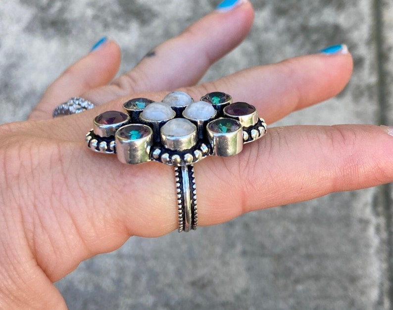May include: A silver ring with a flower-shaped design featuring white moonstone and purple amethyst gemstones.