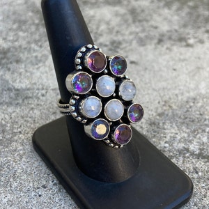 May include: A silver ring with a cluster of round, iridescent gemstones. The ring features a mix of white and purple stones, set in a silver band.