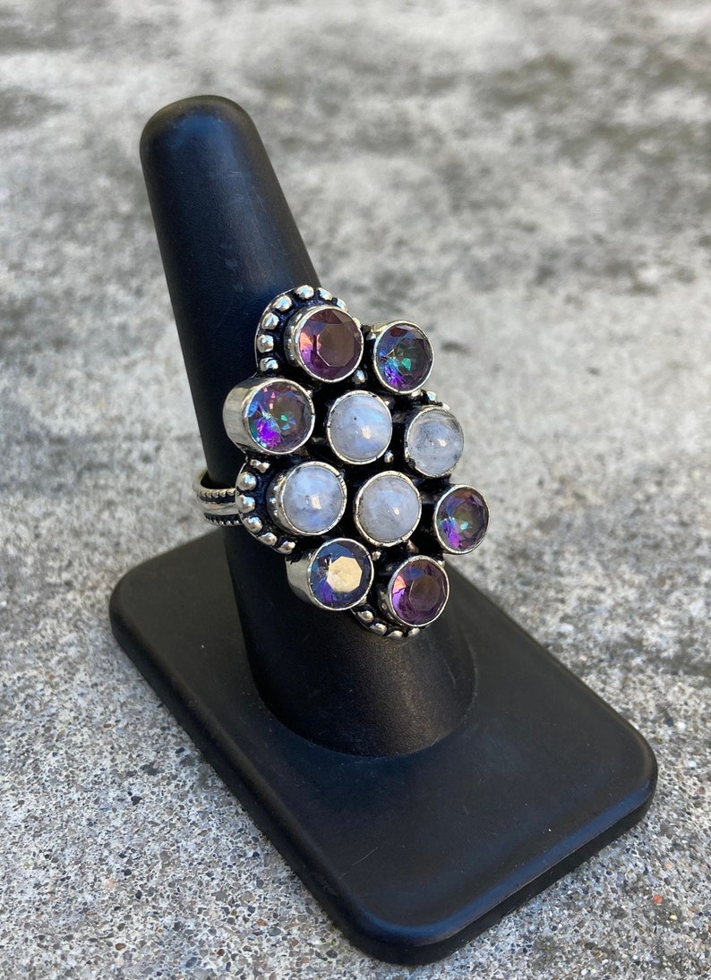 May include: A silver ring with a cluster of round, iridescent gemstones. The ring features a mix of white and purple stones, set in a silver band.