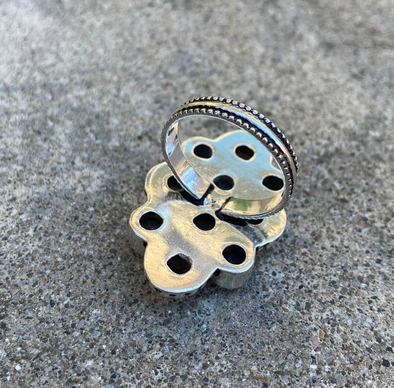 May include: Silver ring with a flower design and multiple small stones. The ring is open on the back and has a beaded edge.