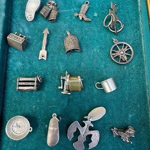 May include: A collection of silver charm pendants in a blue velvet lined box. The charms include a cowboy boot, a Coke bottle, a bell, a wheel, a shoe, a pair of milk bottles, an arrow, a sewing machine, a cup, a plate, a bird, a plane, and a lightning bolt.