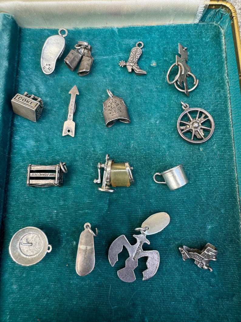 May include: A collection of silver charm pendants in a blue velvet lined box. The charms include a cowboy boot, a Coke bottle, a bell, a wheel, a shoe, a pair of milk bottles, an arrow, a sewing machine, a cup, a plate, a bird, a plane, and a lightning bolt.