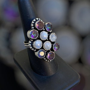 May include: A silver ring with a cluster of iridescent purple and white stones. The ring is set on a black ring holder.