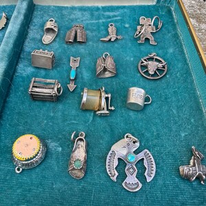 May include: A collection of silver charm bracelets with various designs, including a cowboy boot, a cowboy hat, a baby bottle, a pair of pants, an arrow, a wagon wheel, a cowboy, a horseshoe, a boot, and a bird. The charms are displayed on a blue velvet background.