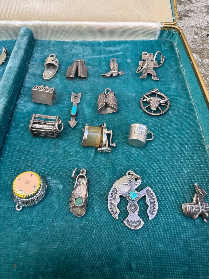 May include: A collection of silver charm bracelets with various designs, including a cowboy boot, a cowboy hat, a baby bottle, a pair of pants, an arrow, a wagon wheel, a cowboy, a horseshoe, a boot, and a bird. The charms are displayed on a blue velvet background.