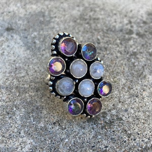 May include: A silver ring with a cluster of round, faceted gemstones. The stones are a mix of purple amethyst, clear moonstone, and iridescent rainbow moonstone.