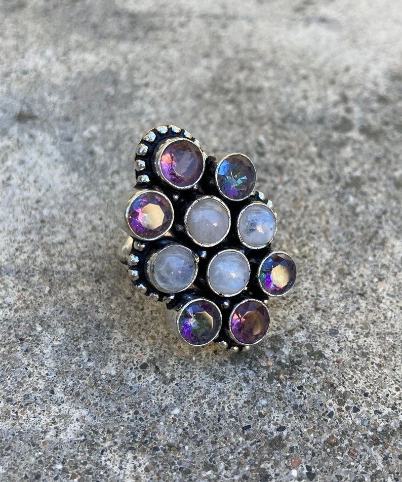 May include: A silver ring with a cluster of round, faceted gemstones. The stones are a mix of purple amethyst, clear moonstone, and iridescent rainbow moonstone.