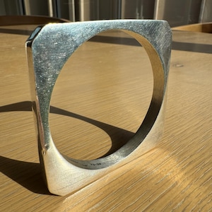 May include: A silver-toned bangle bracelet with a square outer shape and a circular inner opening. The metal surface features a brushed texture and visible markings on the inner edge that read 76-32. The bangle has a thick, solid construction.