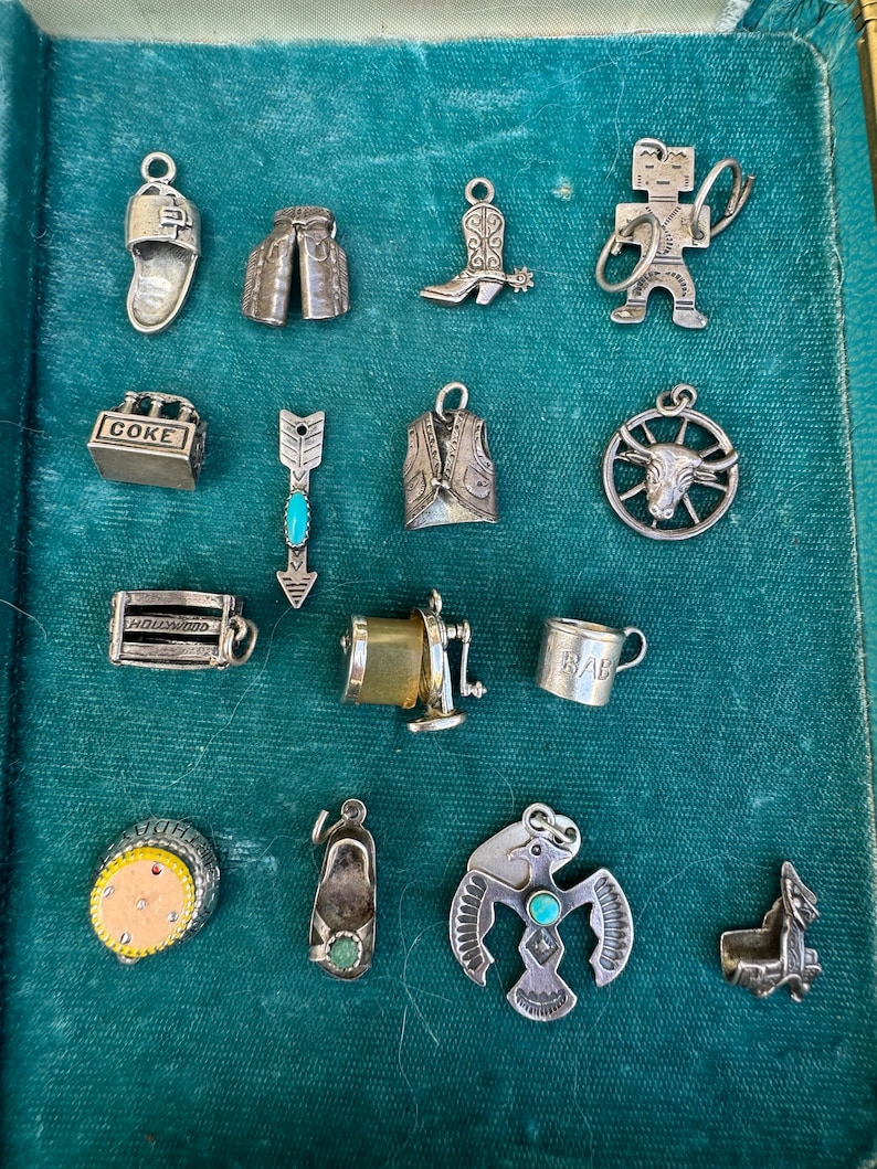 May include: A collection of 14 silver charm pendants, including a cowboy boot, a vest, a Native American headdress, a Coke bottle, a baby cup, a pair of cowboy chaps, a pair of slippers, a Hollywood sign, a wheel with a bull's head, an arrow with a turquoise stone, a Native American figure, and a pair of slippers.