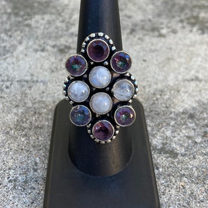 May include: A silver ring with a cluster of moonstone and amethyst gemstones. The ring is set in a decorative, ornate design.