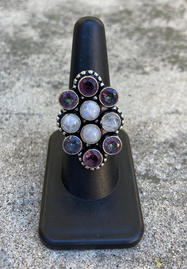 May include: A silver ring with a cluster of moonstone and amethyst gemstones. The ring is set in a decorative, ornate design.