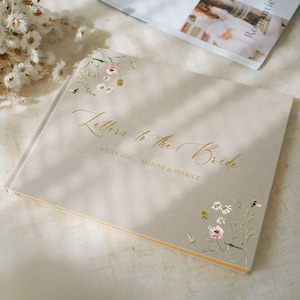 May include: A beige wedding guest book with gold script reading "Letters to the Bride". The cover features floral illustrations and the date "6 juin 2016". A bouquet of white flowers is in the background.