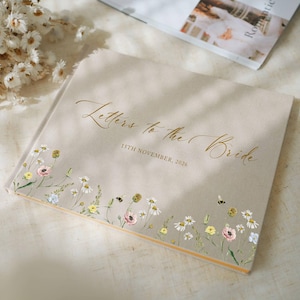 Puede incluir: Un libro de visitas de boda beige titulado "Letters to the Bride" con letras doradas. El libro está decorado con un borde floral de margaritas, amapolas y otras flores silvestres. La fecha "15 de noviembre de 2026" está impresa debajo.