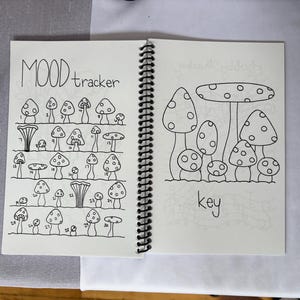 May include: A spiral-bound notebook with black ink illustrations of mushrooms. The left page has a mood tracker with numbered mushroom drawings. The right page has a key with mushroom drawings. The word "mood" and "tracker" are written at the top of the left page.