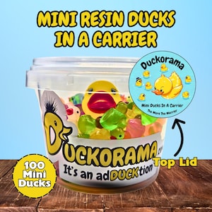 May include: A clear plastic container filled with mini resin ducks and colorful jelly candies. The container has a lid and a label that reads "Duckorama" and "100 Mini Ducks". The text "Mini Resin Ducks In A Carrier" is at the top.