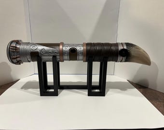 May include: A silver and brown lightsaber with a unique design, featuring a curved handle and a black leather wrap. The lightsaber is displayed on a black stand.